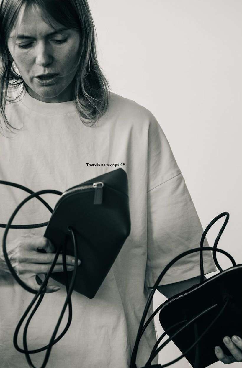 Monochromatic portrait of an adult woman holding two black leather handbags with cord straps against a white background in a studio.