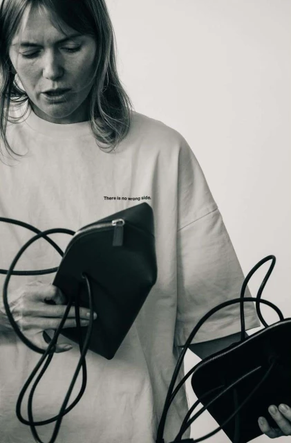 Monochromatic portrait of an adult woman holding two black leather handbags with cord straps against a white background in a studio.