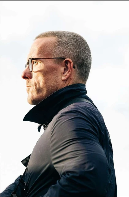 Profile portrait of an adult man with glasses, wearing a jacket, set against a bright sky. The man is looking off to the side.