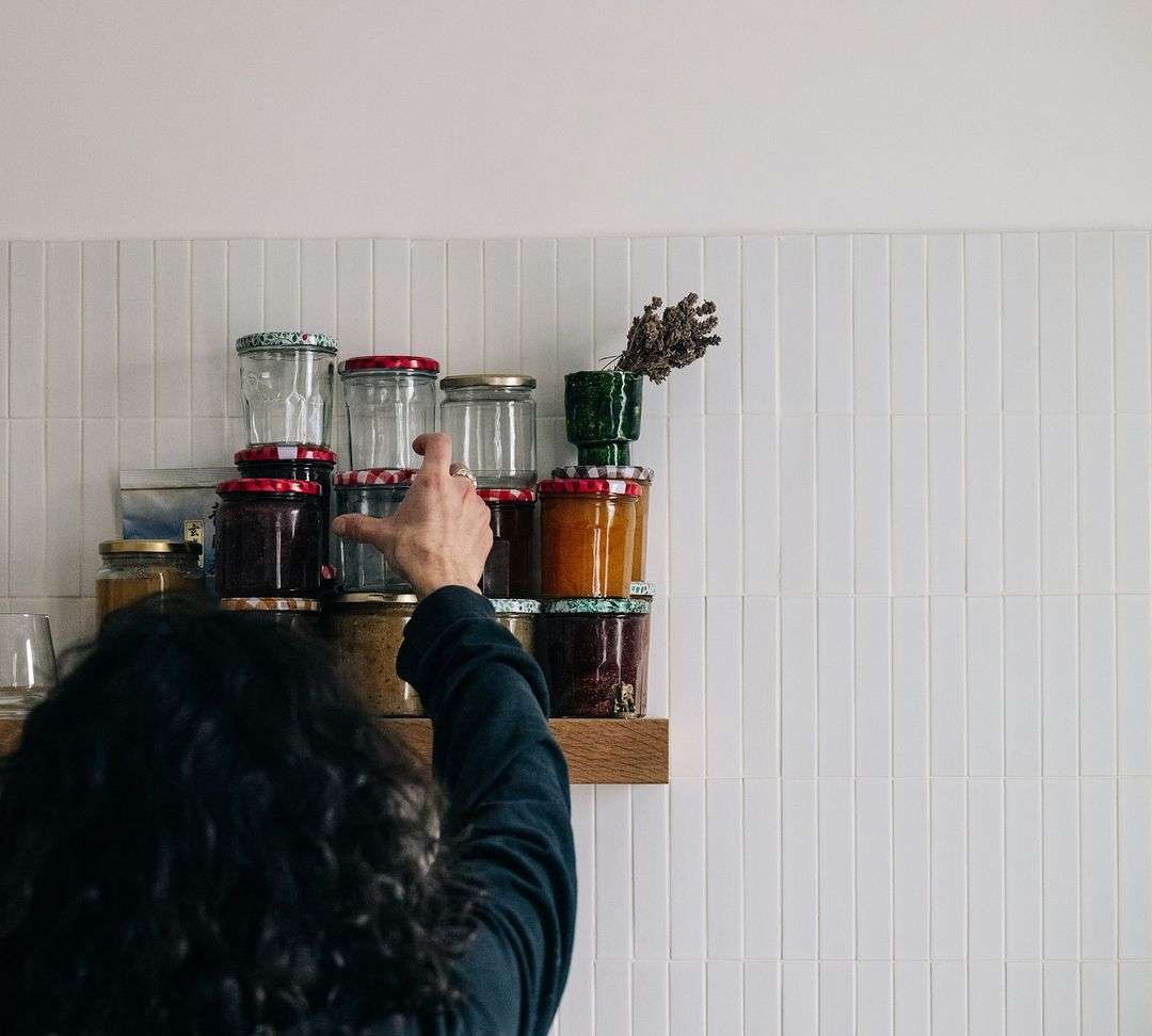A woman reaches for a glass jar on a shelf filled with jars of preserved goods, set against a white tiled wall in a well-lit kitchen.