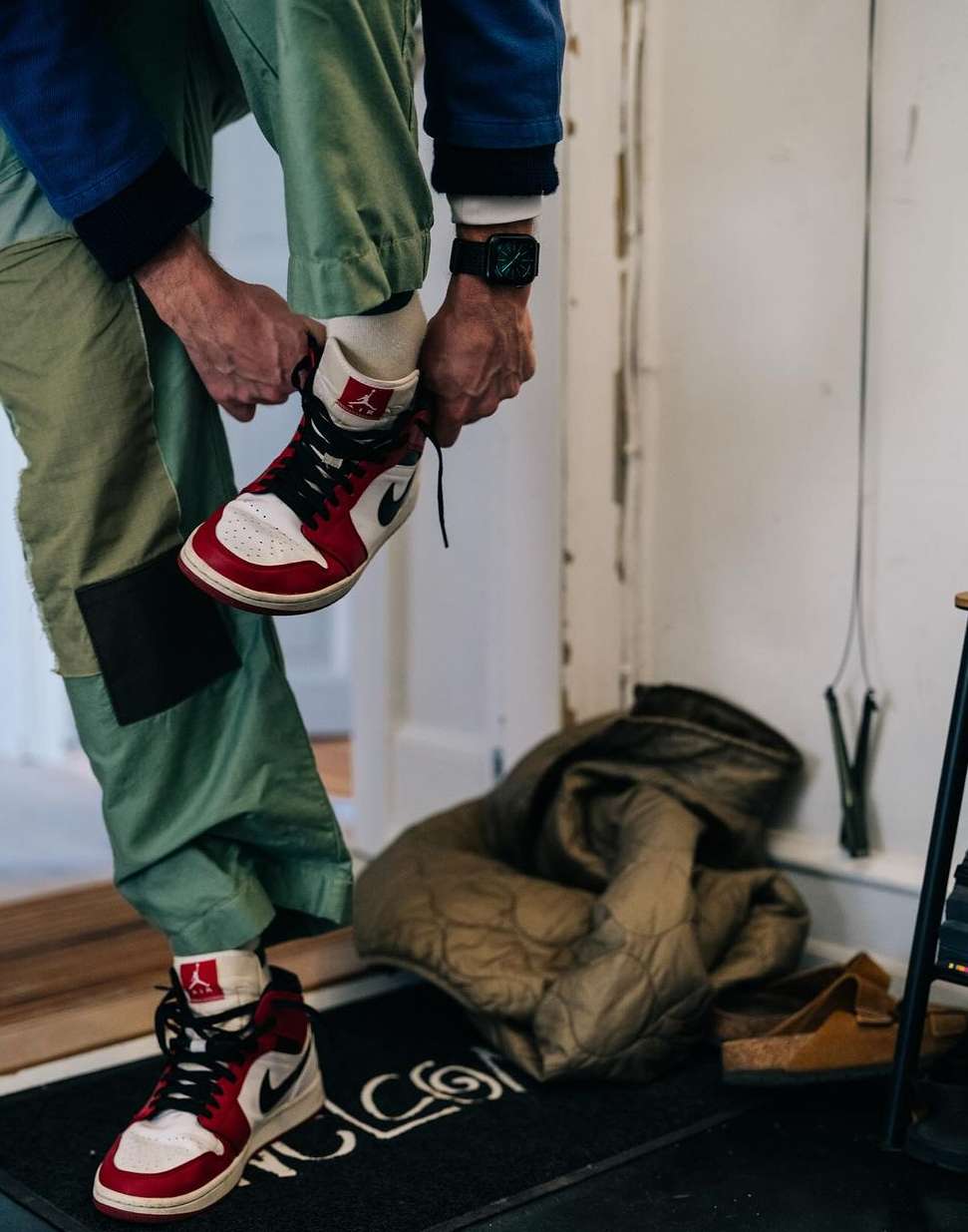 Close-up of a person tying red, white, and black high-top Nike Air Jordan 1 sneakers in an indoor setting with a welcome mat.