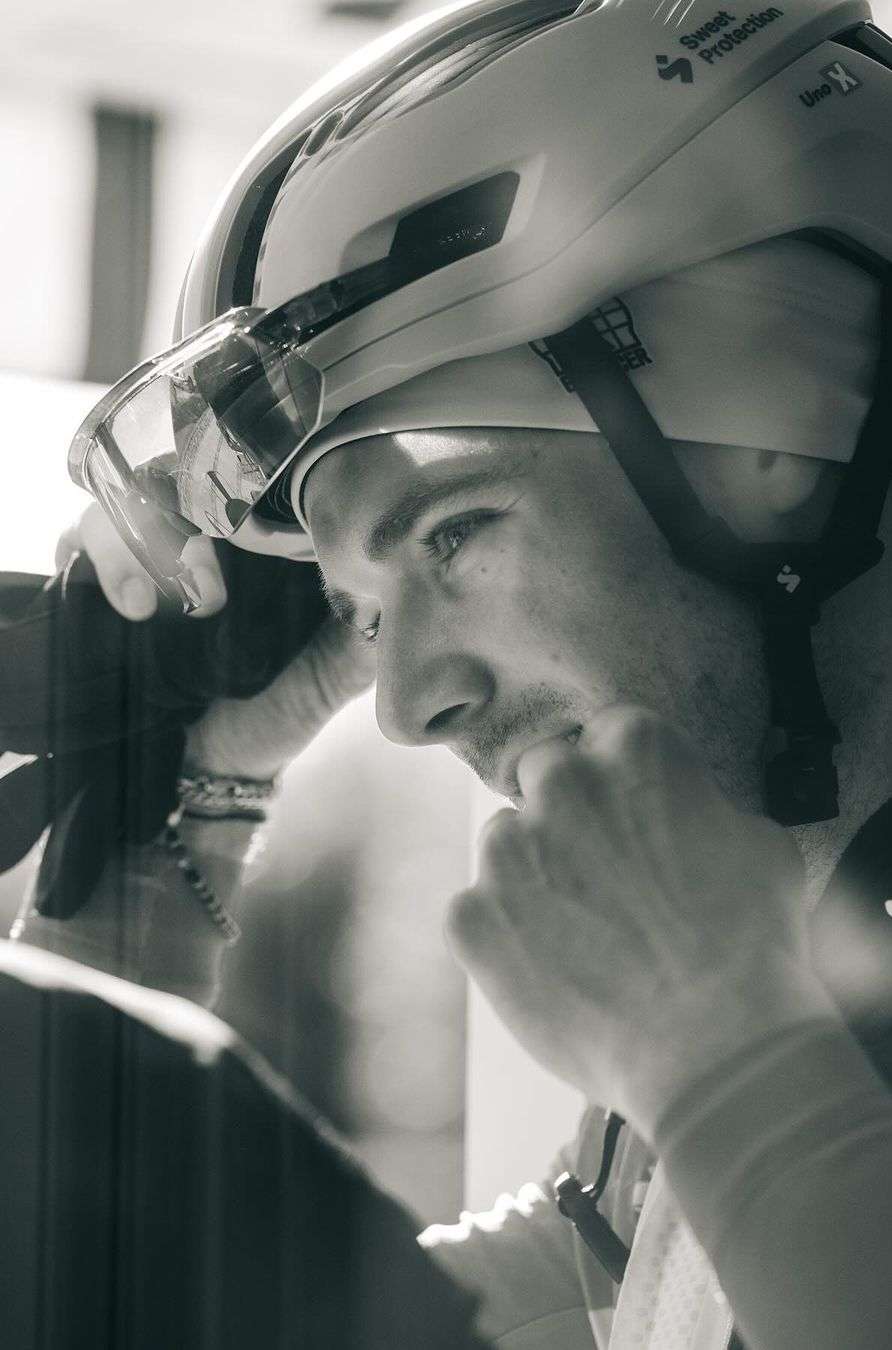 Monochrome portrait of a man wearing a cycling helmet with visor and adjusting the straps, indoor environment, close-up framing.