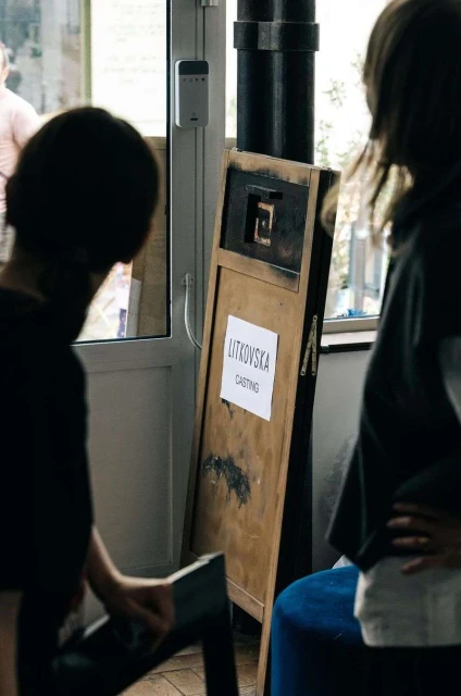 Two people stand near a wooden board displaying 'Litkovska Casting' in an indoor setting with a white door and stool.