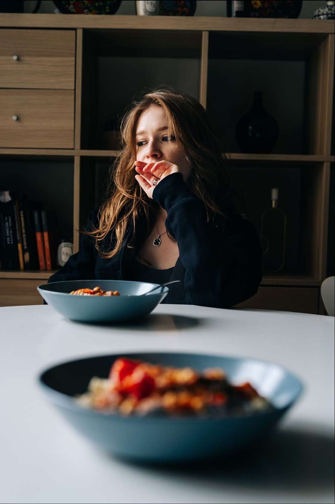 A woman sits at a table with two bowls of food in a dimly lit room with a light wood shelf and books in the background.