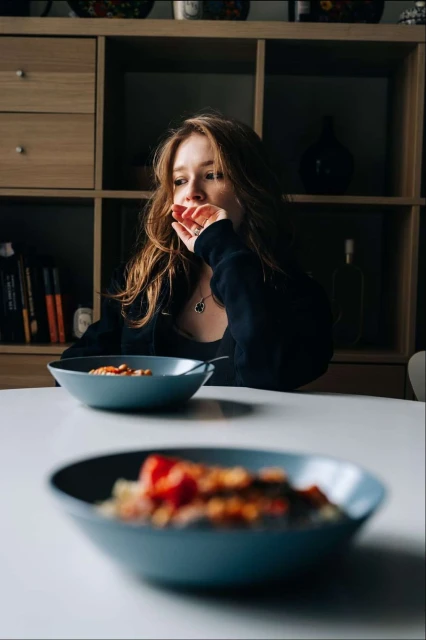 A woman sits at a table with two bowls of food in a dimly lit room with a light wood shelf and books in the background.