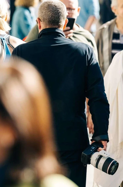 Back view of a man with short hair in a black jacket holding a digital camera amongst a blurred crowd outdoors in daytime.