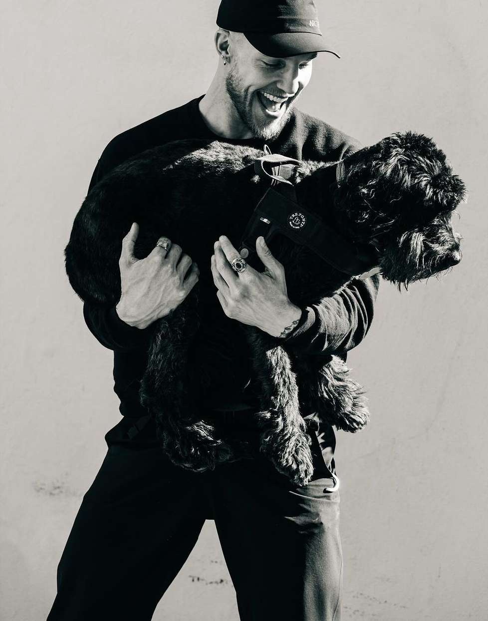 Happy adult male smiles and holds a black dog outdoors in a black and white photograph against a neutral background with soft lighting.