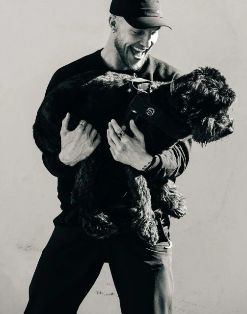 Happy adult male smiles and holds a black dog outdoors in a black and white photograph against a neutral background with soft lighting.
