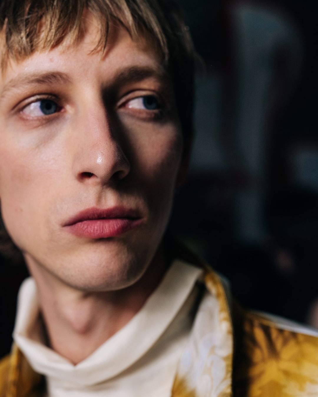 Close-up portrait of a male model with light hair and blue eyes wearing a patterned jacket and white turtleneck.