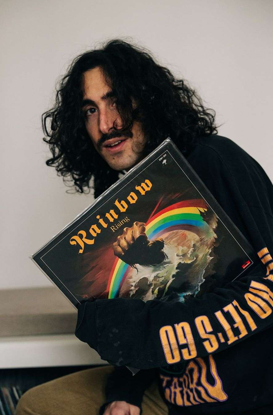 Young adult poses indoors holding Rainbow Rising vinyl record album, wearing black long sleeve shirt with gold text on sleeve.