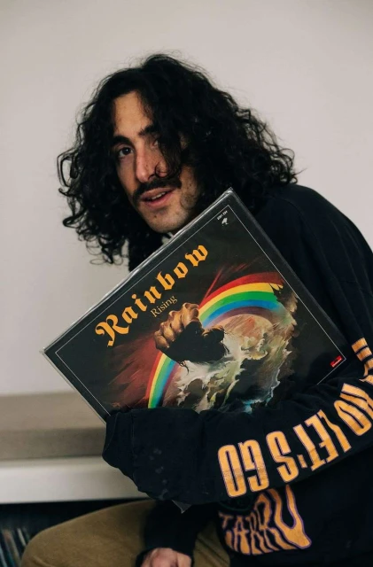 Young adult poses indoors holding Rainbow Rising vinyl record album, wearing black long sleeve shirt with gold text on sleeve.