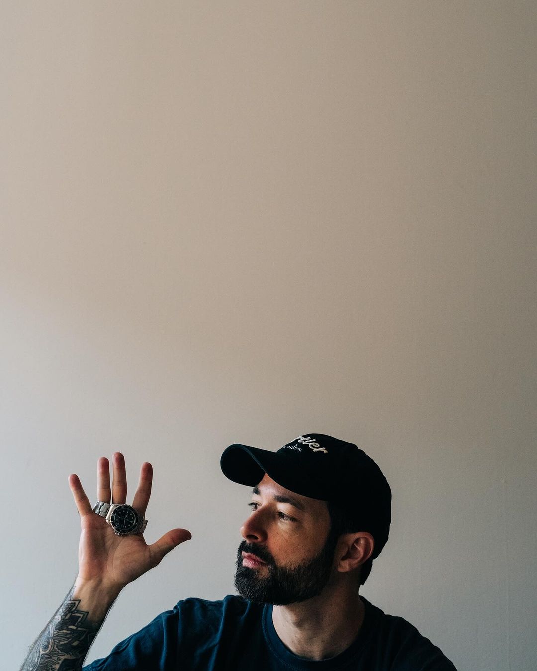 Man wearing a dark baseball cap and t-shirt displaying a wristwatch on his raised open palm, photographed by Adam Katz Sinding