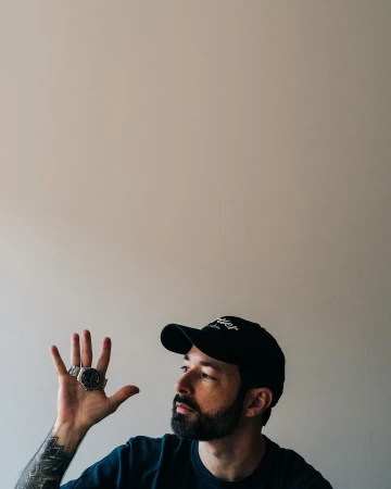 Man wearing a dark baseball cap and t-shirt displaying a wristwatch on his raised open palm, photographed by Adam Katz Sinding