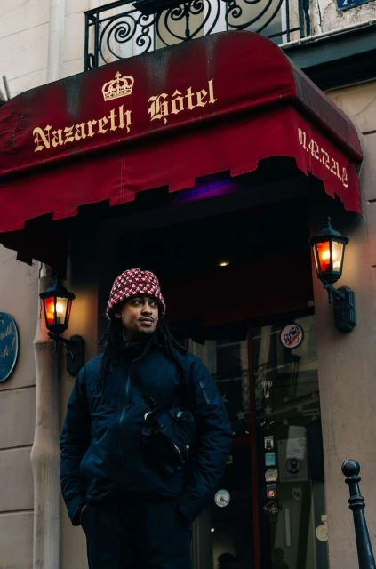 Man stands in front of the Nazareth Hôtel in Paris wearing a dark jacket and patterned hat. Street lamps flank the entrance.