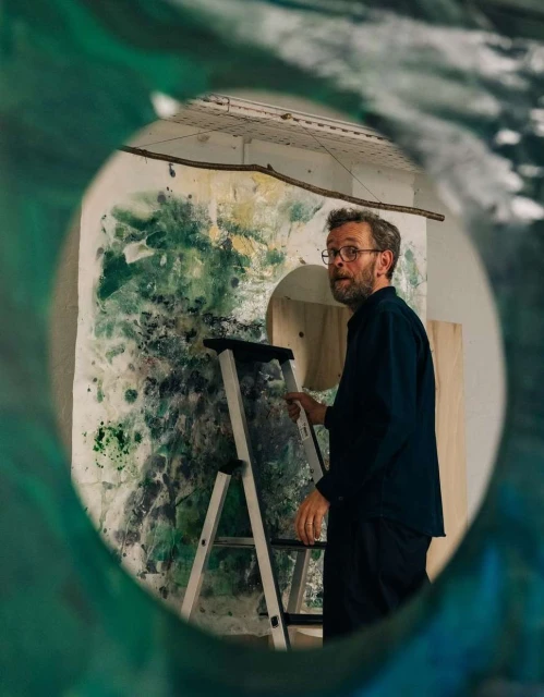 Man with glasses stands on a ladder next to an abstract painting in an art studio, viewed through a round shape, indoor setting, daylight.