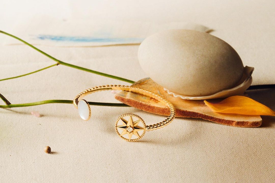 Elegant golden bangle with a star motif and white accents, artfully presented with a seashell and floral elements in a neutral-toned setting.