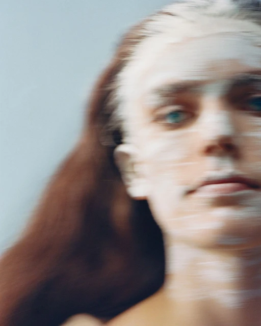 Close-up of an adult woman with pale face paint and blue eyes, blurred to convey motion; her auburn hair is visible against a pale blue backdrop.