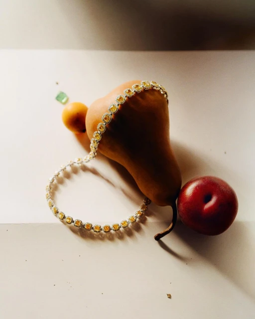 A delicate yellow gemstone bracelet encircles a brown pear and a red plum on a white surface, creating an elegant jewelry still life.