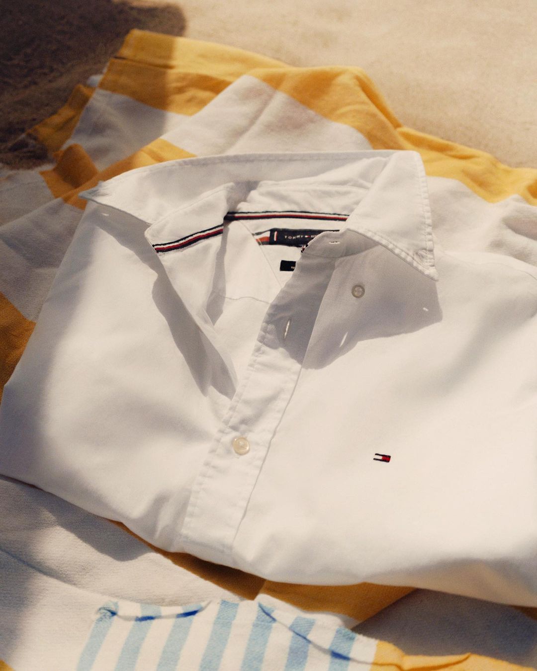 White Tommy Hilfiger button-down shirt laid flat on a striped blanket in sunlight, showcasing the brand's design and quality.