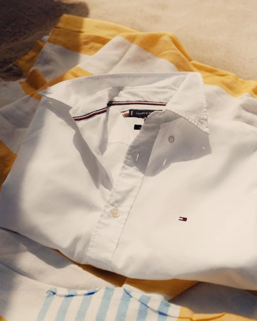 White Tommy Hilfiger button-down shirt laid flat on a striped blanket in sunlight, showcasing the brand's design and quality.