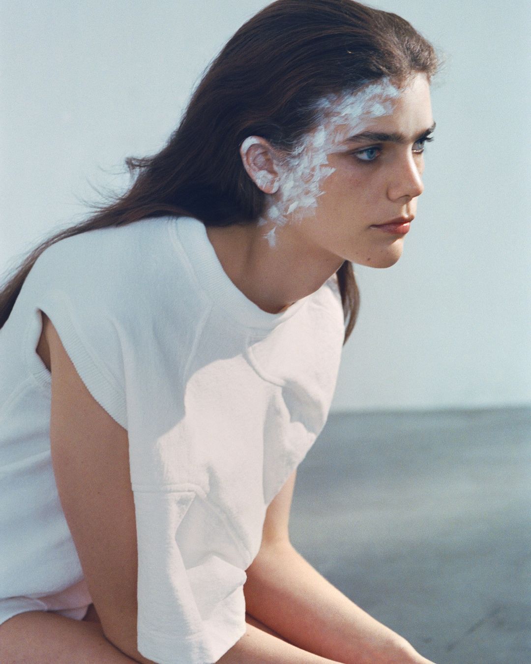 Portrait of a woman with white face paint on her temple and ear, wearing a white t shirt against a neutral studio background.