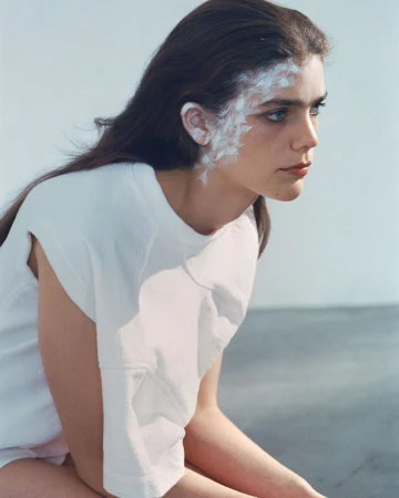Portrait of a woman with white face paint on her temple and ear, wearing a white t shirt against a neutral studio background.