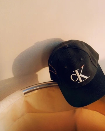 A black baseball cap with white CK logo resting near a wall casting a shadow, photographed by Adam Kremer.