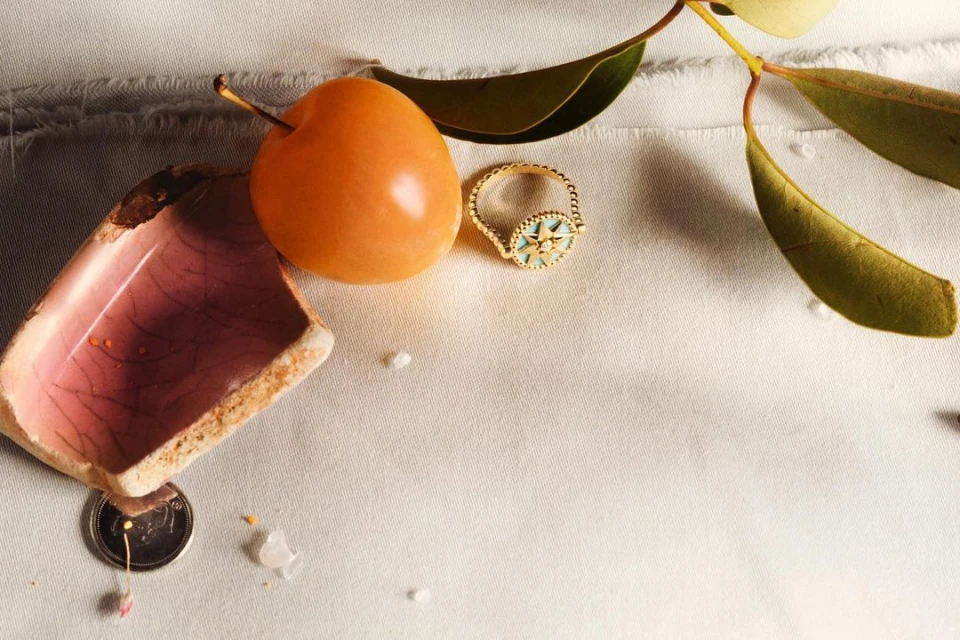 Still life captures a gold ring, orange fruit, broken ceramic, and leaves on a textured white surface under soft lighting.
