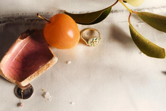 Still life captures a gold ring, orange fruit, broken ceramic, and leaves on a textured white surface under soft lighting.