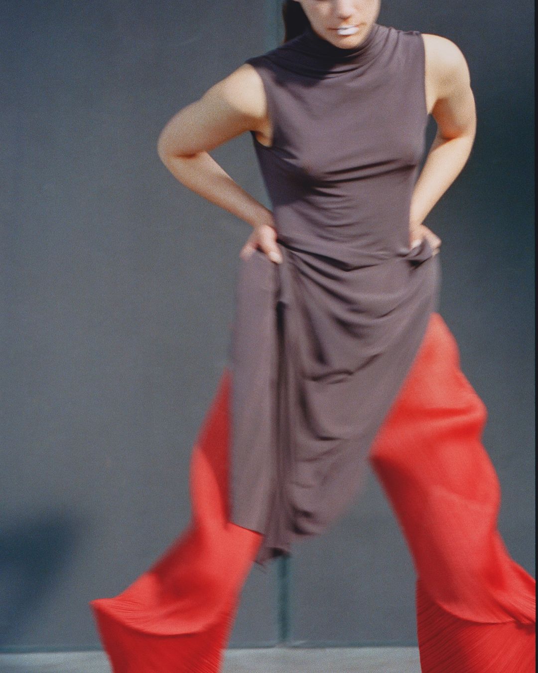 A woman in a taupe dress and red pants strikes a pose against a neutral backdrop, showcasing a modern fashion aesthetic with a studio vibe.