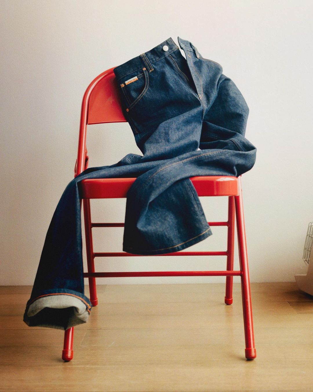 Dark wash denim jeans are draped over a red metal chair in a minimalist, light-filled photo studio setting with a wood floor.