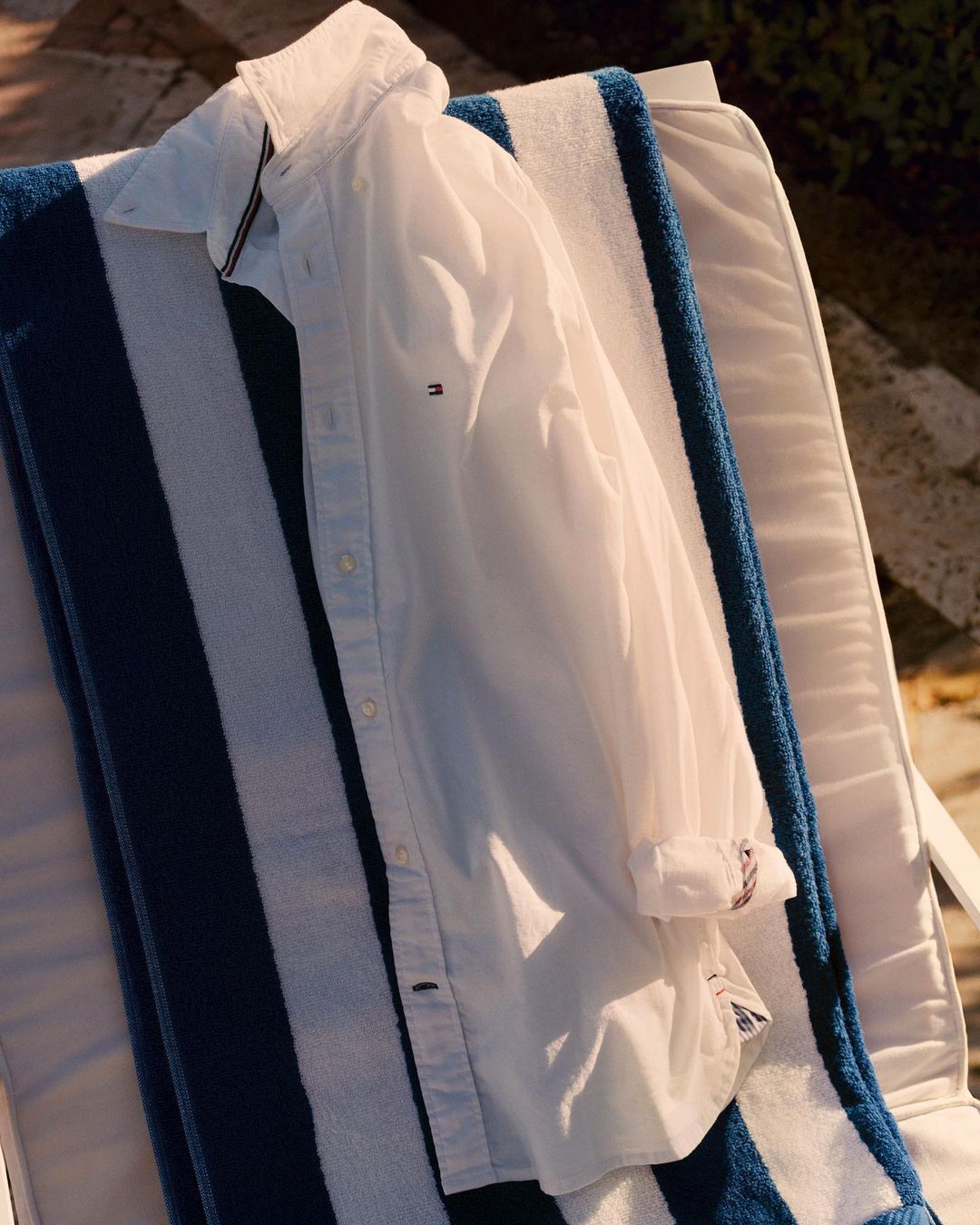 White button-down shirt sits on a blue and white striped beach towel draped over a white deck chair in outdoor setting.