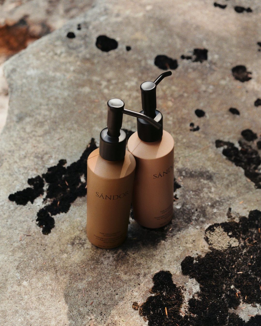 Two tan pump bottles with black pumps stand on a textured gray stone surface with dark spots, photographed in a product shot.