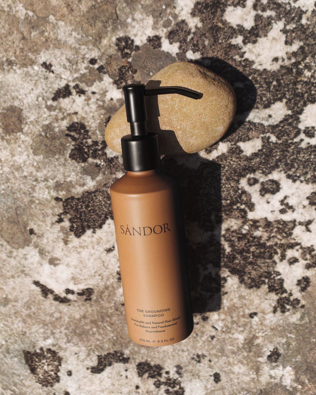 A tan bottle of Sandor shampoo with a black pump sits against a rock, accented by a small rock in the background.