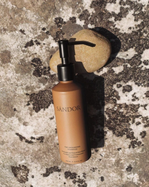 A tan bottle of Sandor shampoo with a black pump sits against a rock, accented by a small rock in the background.