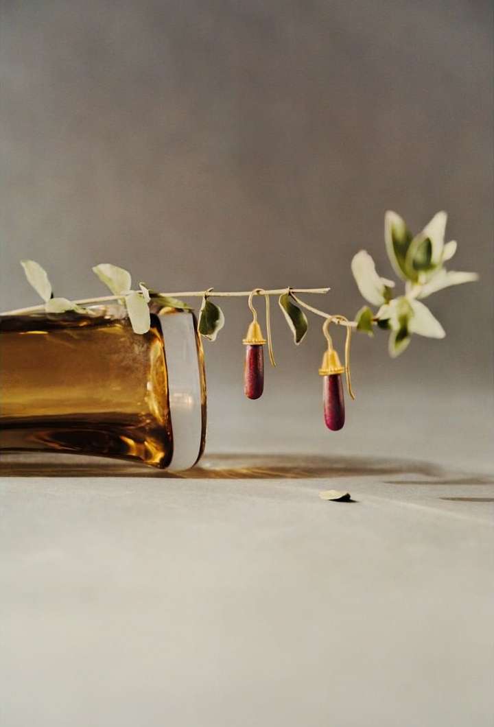 Elegant still life featuring two red pendant earrings hanging from a leafy branch next to a gold-colored glass bottle on a table.