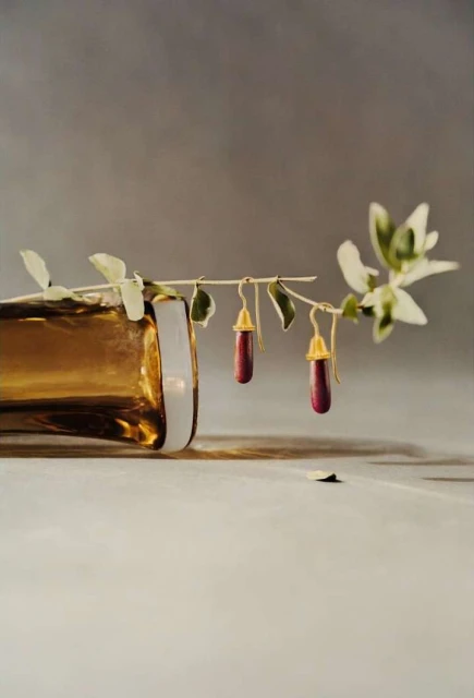 Elegant still life featuring two red pendant earrings hanging from a leafy branch next to a gold-colored glass bottle on a table.