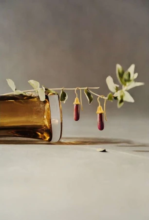 Elegant still life featuring two red pendant earrings hanging from a leafy branch next to a gold-colored glass bottle on a table.