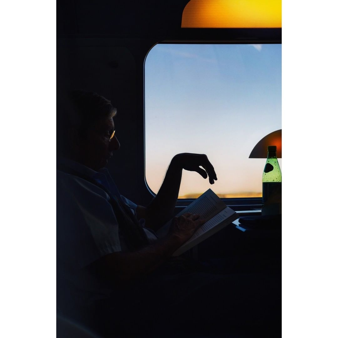 Silhouetted man reads a book on a train at sunset with a lamp and bottled water nearby, creating a serene and cozy travel scene.