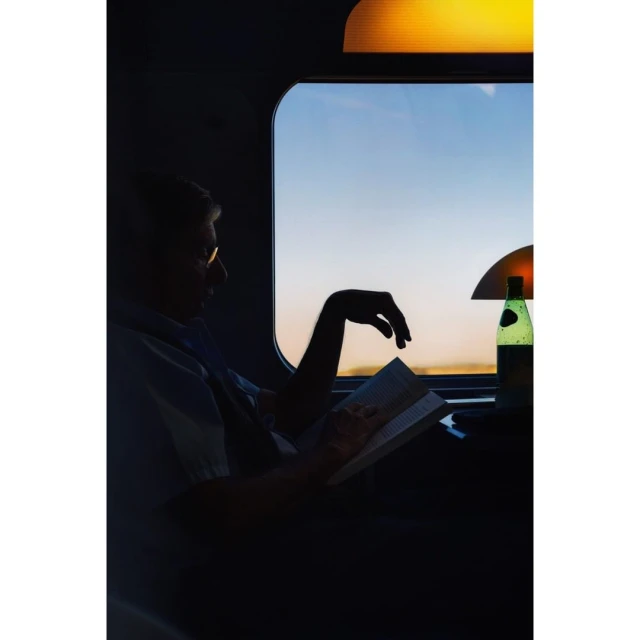 Silhouetted man reads a book on a train at sunset with a lamp and bottled water nearby, creating a serene and cozy travel scene.