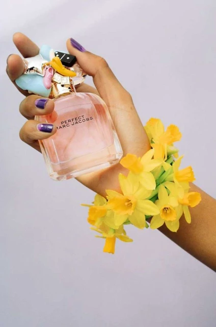 Woman's hand holding Perfect Marc Jacobs perfume bottle adorned with flowers. Close-up studio shot against a white backdrop.