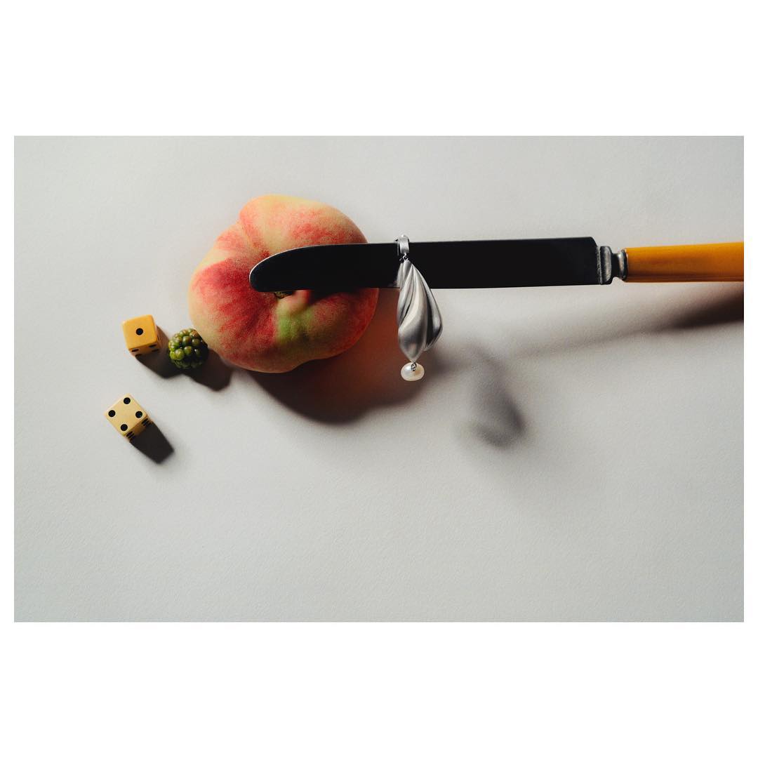 Still life of a peach cut by a knife, with dice, green berries, and silver pendant on a white surface. Studio lighting.