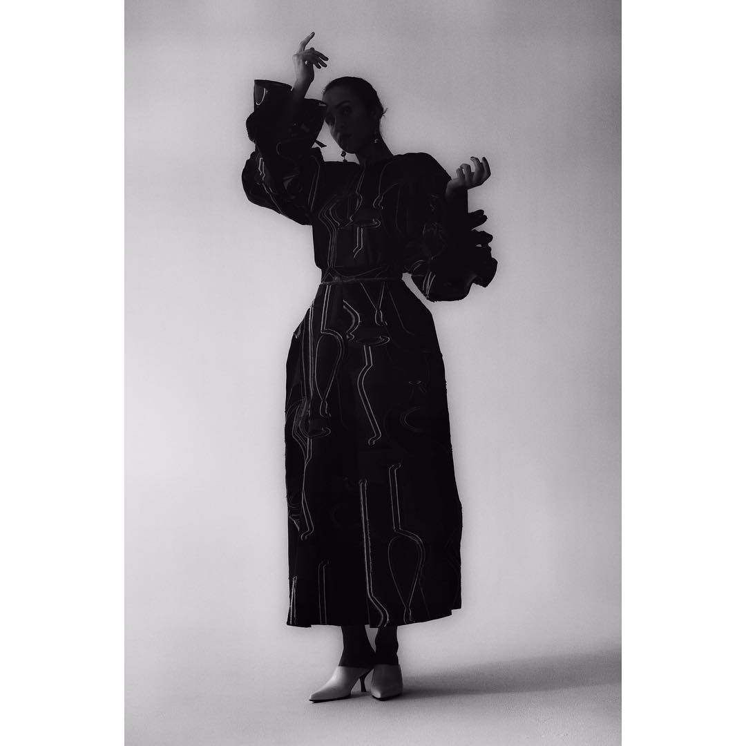 A woman models a dark, oversized dress with white patterns against a stark white background in a high-contrast, black-and-white studio shot.