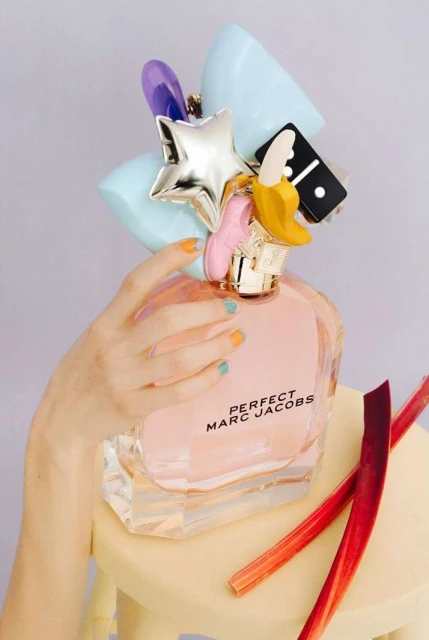 Perfume bottle with whimsical charms on a yellow stool, set against a light purple background, with a hand and rhubarb sticks nearby.