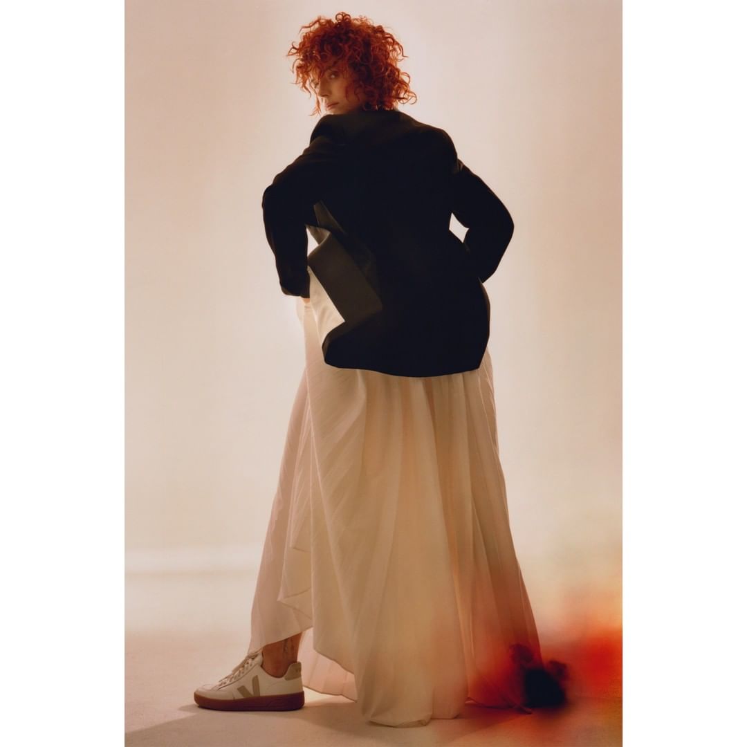 Adult woman with curly auburn hair in black blazer sitting backward on chair with long white skirt, facing away from the camera.