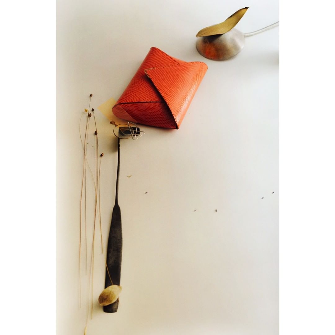 Still life composition featuring an orange clutch, a metal scoop with a leaf, and thin stalks of grass, arranged on a white surface.