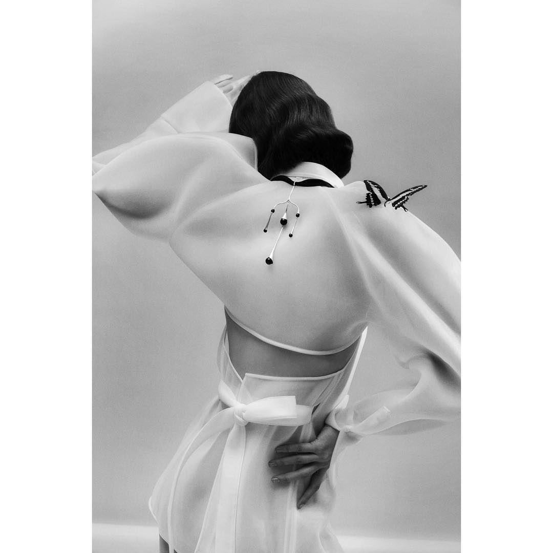 Back view of an adult female with dark, shoulder-length wavy hair. She is wearing a sheer, short white dress with a large butterfly.