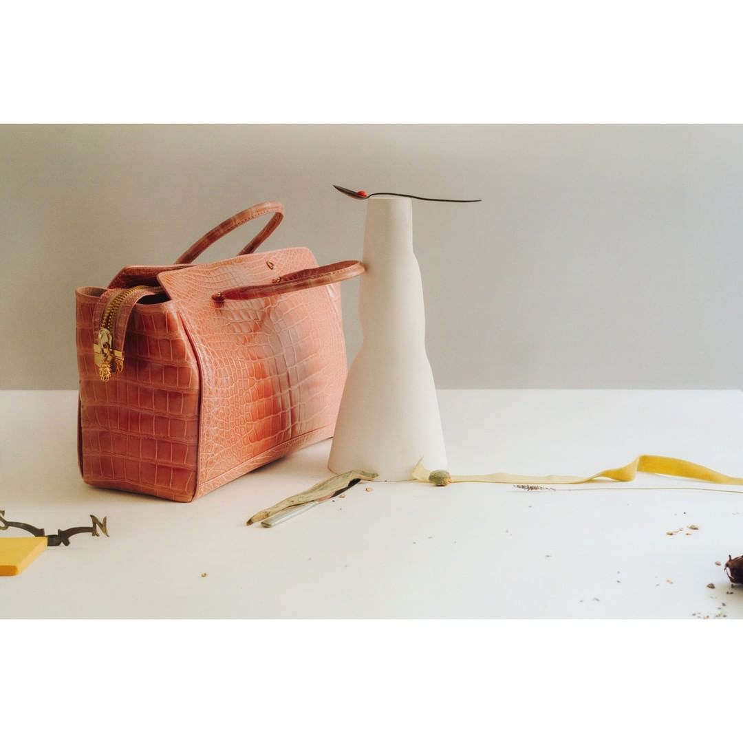 Studio shot of a salmon-colored purse next to a white vase with scattered debris and yellow ribbon on a white surface against a grey background.