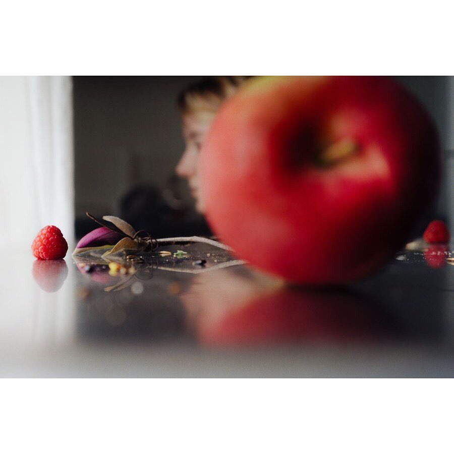 Close-up of a glossy black table with an apple and a raspberry in focus, a blurred profile of a fair-skinned human in background.
