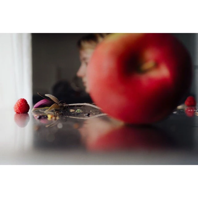 Close-up of a glossy black table with an apple and a raspberry in focus, a blurred profile of a fair-skinned human in background.